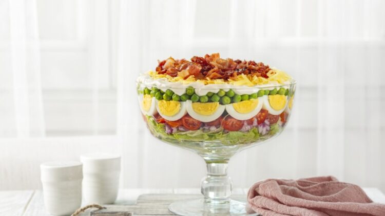 Seven-layer salad. American dish with ingredients: iceberg lettuce, tomatoes, red onions, sweet peas, hard boiled eggs, cheddar cheese, and bacon pieces topped with a mayonnaise dressing.
