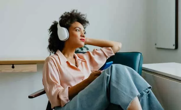 A smiling Afro woman sitting comfortably in an armchair with her earphones on, listening to music on her smartphone
