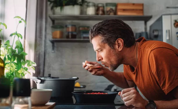 Apprenez à faire la cuisine 1 - Le Progrès Egyptien Close up shot of a handsome young happy Caucasian man tasting sauce with a mixing spoon with his eyes closed over a frying pan in a kitchen.