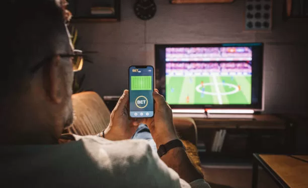 Mature men sitting on sofa at home and watching soccer game.  He is using smart phone for sports betting .